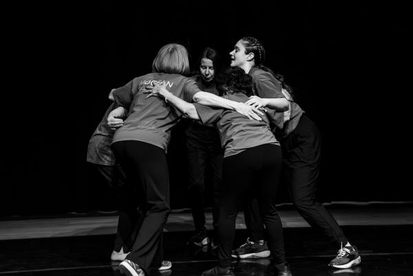 a group of older adults in blue t-shirts stand in a circle with arms embraced on a dark stage