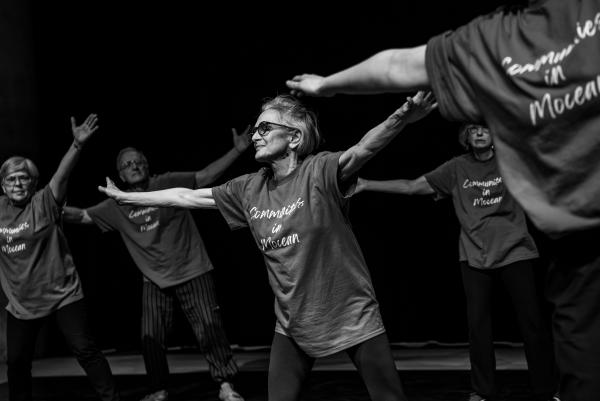 a black an white photography of a  group of older adults in blue t-shirts stand on a stage with arms outstretched in a V