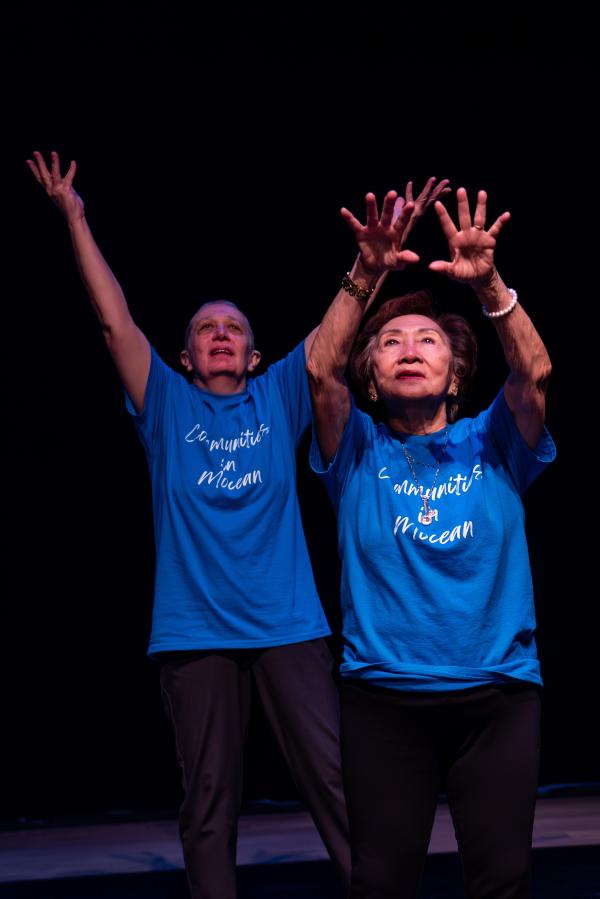 Elderly dancer in blue shirt against black background, arms reaching