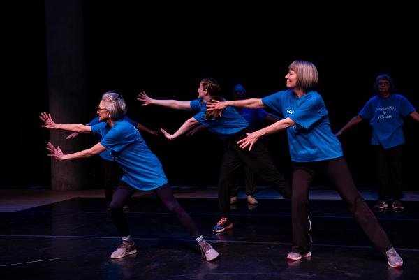 Elderly dancer in blue shirt against black background, arms reaching
