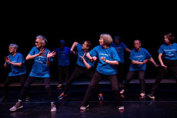 Group of elderly dancers in blue shirts embrace in a circle against black background