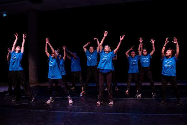 Elderly dancers in blue shirts against black background, arms reaching