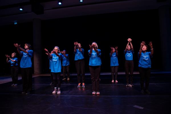Elderly dancers in blue shirts against black background, arms reaching