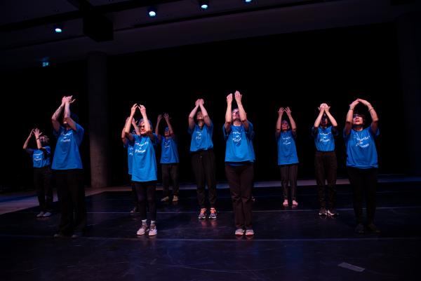 Elderly dancers in blue shirts against black background, arms reaching
