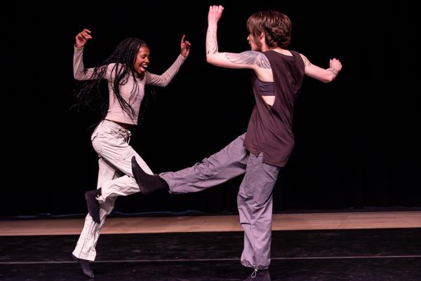 One black and one white female dancers face each other with arms in the air and hopping.