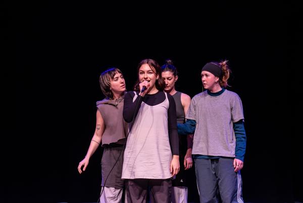 Three white dancers look at the center dancer with a microphone