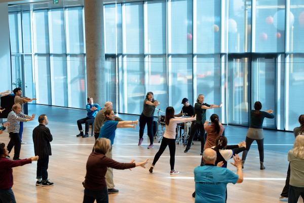 In a bright room full of large floor to ceiling windows a group of diverse people strtch to the right together in a group movement class centered around a percussionist and lead teacher wearing a shirt that says Mocean Dance. Some of the participants are seated and others standing.  