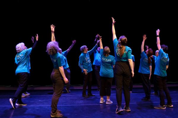 A group of intergenerational dancers in a circle reach one arm up