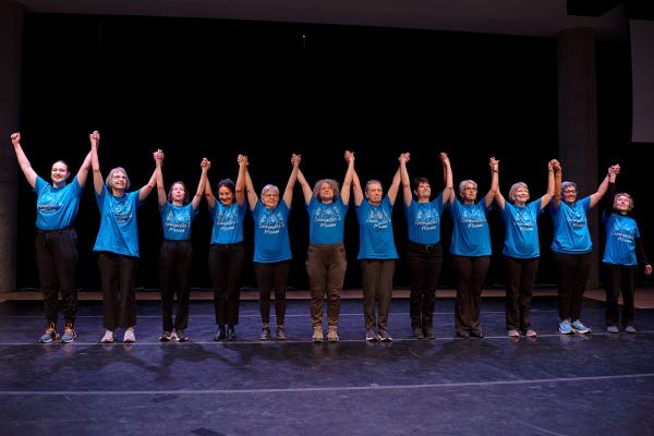 Senior dancers in blue t-shirts hold hands in a line with their arms straight up