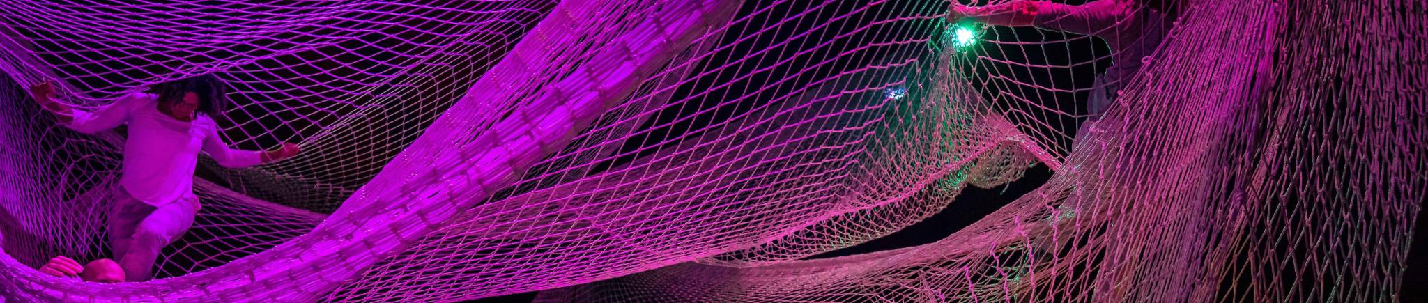Two people stand or sit within layers of suspended nettings lit in purple.