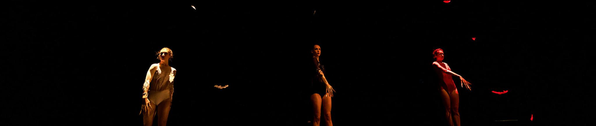 Three dancers stand in a dark space in their own spotlights.
