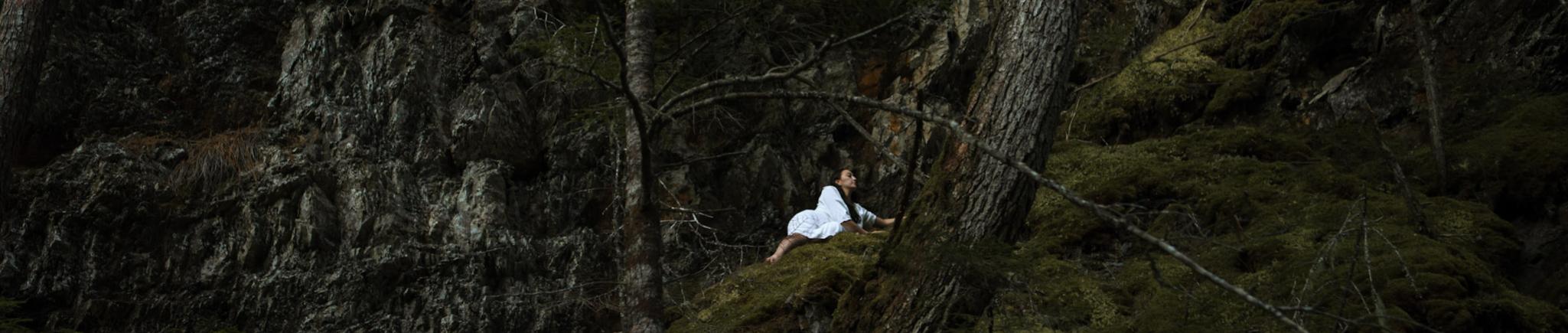 A woman wearing white lays down in a dark forest.