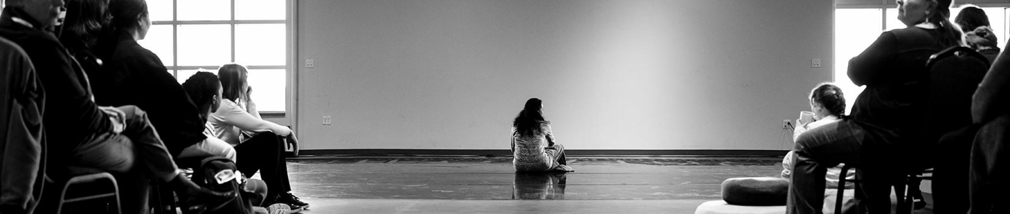 A woman sits on the floor between two banks of seated viewers..