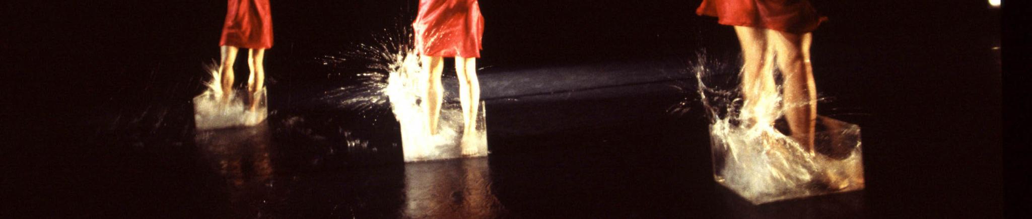 Three dancers stand in clear buckets filled with splashing water. 