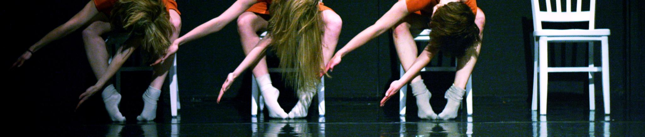 Three dancers pose leaning forward on white chairs.