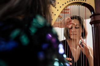 Over a persons shoulder, a person can be seen through the strings of a harp, as many hands pluck the strings.