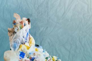 A woman stands shrouded in plastic debris over a blue textured background.