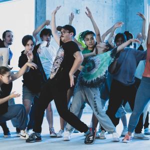 A group of people dance with their arms raised in front of a concrete wall with windows.