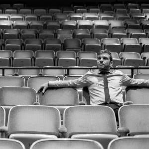 A black and white image of a man in a shirt and tie sitting in an empty theatre.