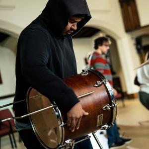 A man in a hoodie holds a drum sideways. People dance in the background.