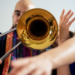 A person plays the trombone while another person reaches arms around the bell of the instrument 