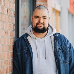 A man in a jean jacket and hoodie smiles next to a brick wall.