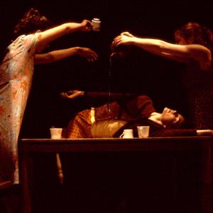 Two dancers pour tea on a dancer laying on a wooden table. 