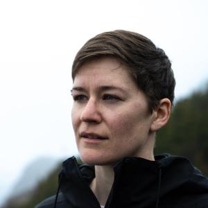 A woman stands in front of a steep forested coastline