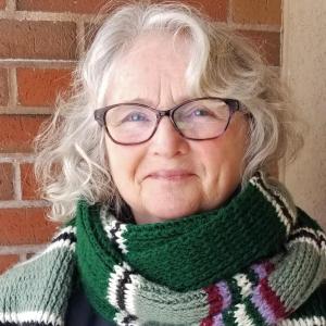 A white-haired woman in glasses and a striped scarf smiles.