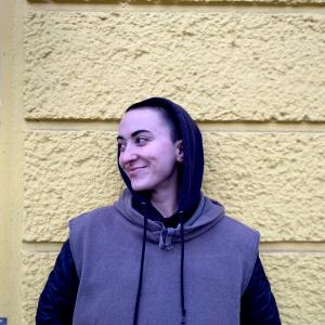 A person with short hair in a hoodie stands in front of yellow wall.