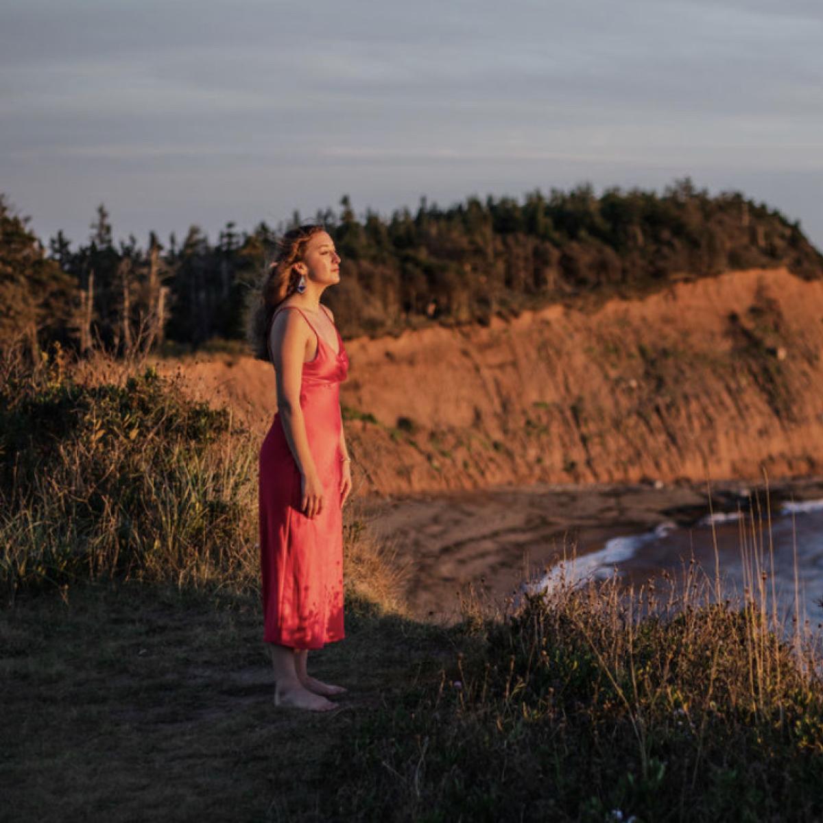 A young blonde woman in a bright red satin dress stands on a red sandy cliff overlooking the ocean.