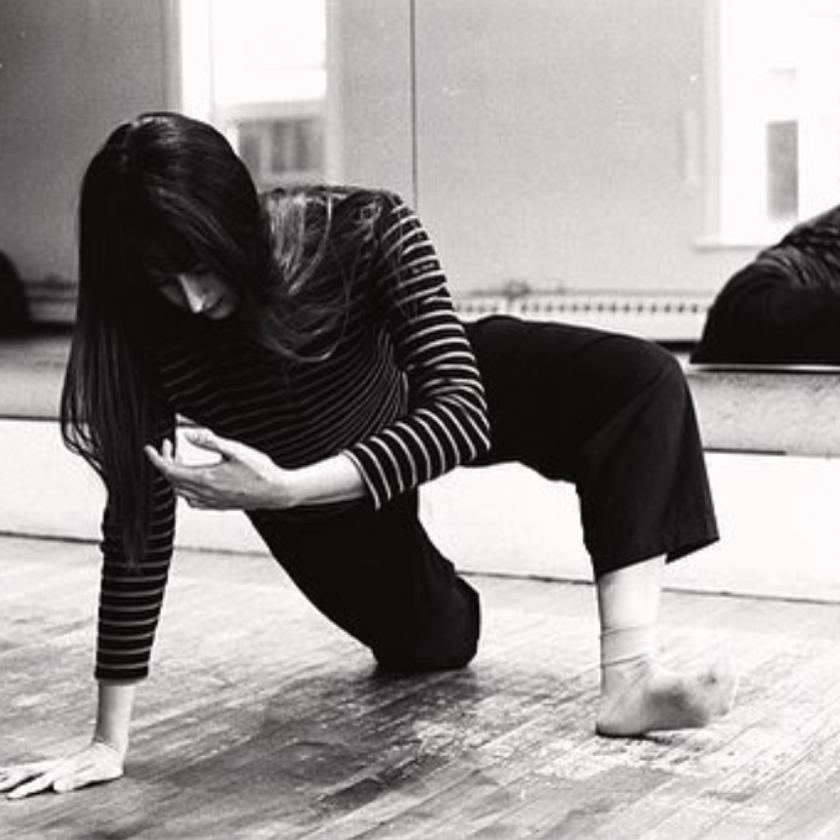 Black and white image of Lauren in a bright dance studio. They have long hair and are dressed in stripes. They are peeling themselves up from the ground which you also see in the mirror behind them.
