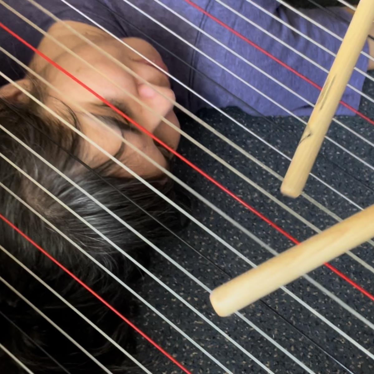 A woman lays under the strings of a harp, with drum sticks laying on top.