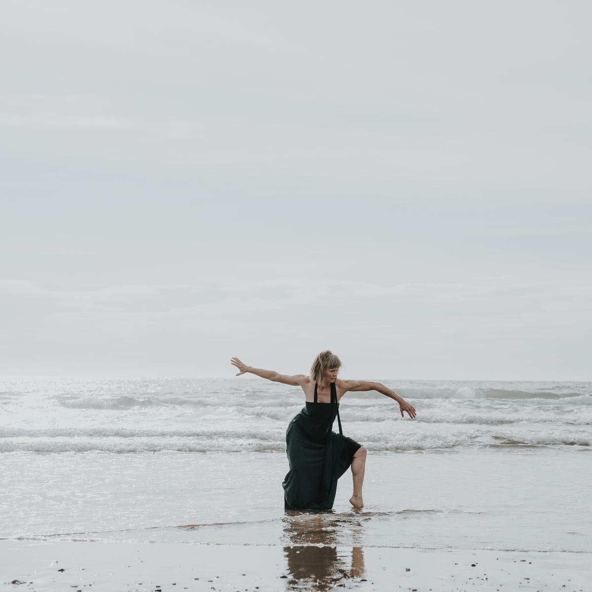 A woman in a black dress dances in the ocean waves on the beach