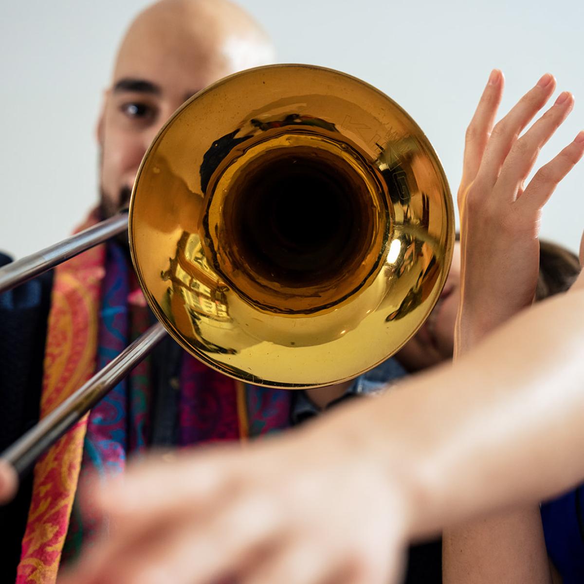 A person plays a trombone while another reaches soft hands around the bell of the instrument.