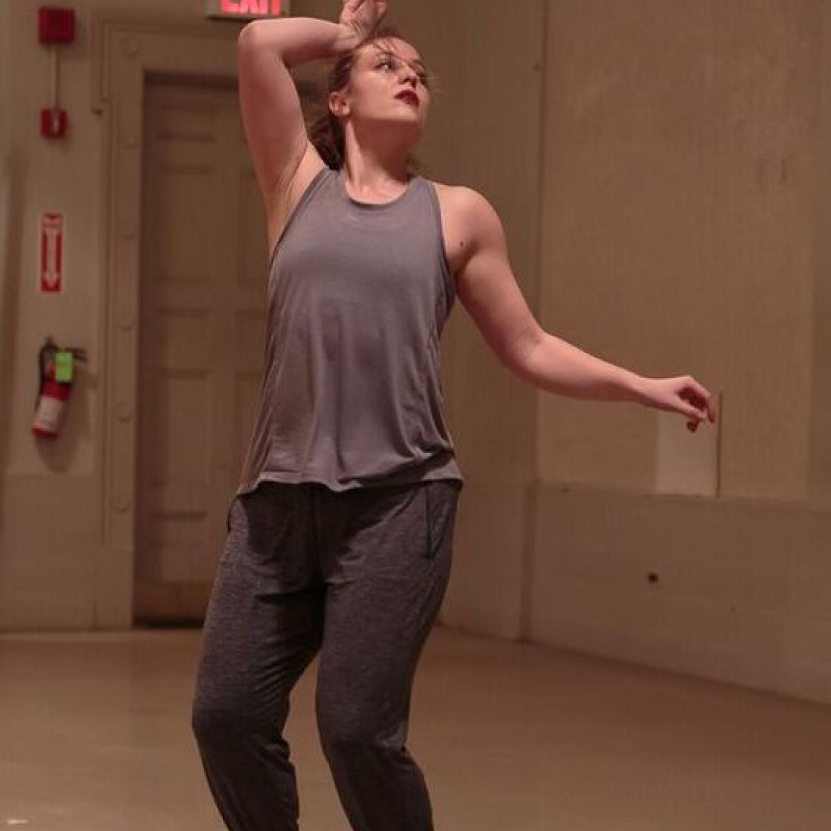 A white women in a pale studio in a beige tank top stands on demi-pointe, knees bent with her right wrist at the forehead. 
