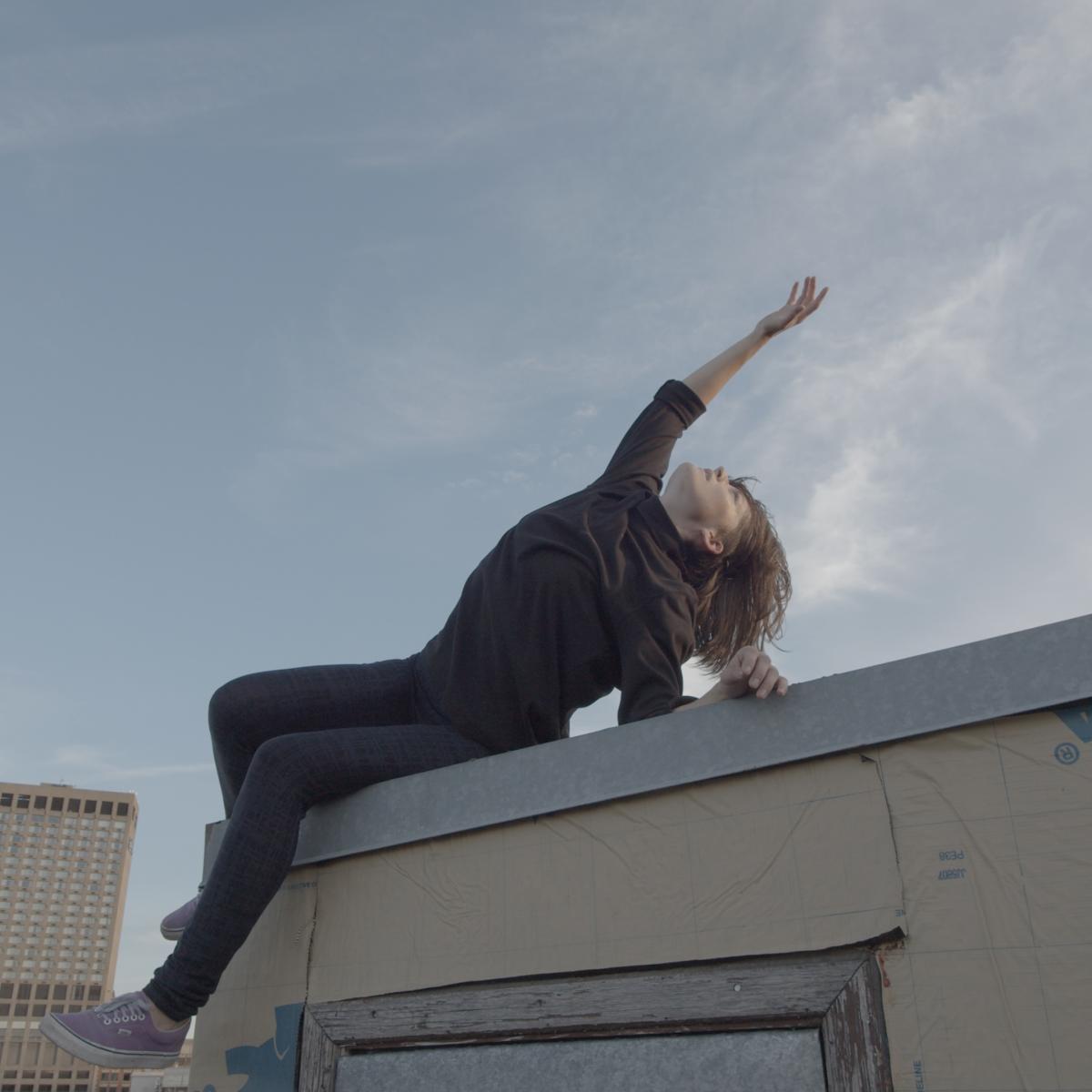 A woman in black sits on the edge of a roof reaching to the sky