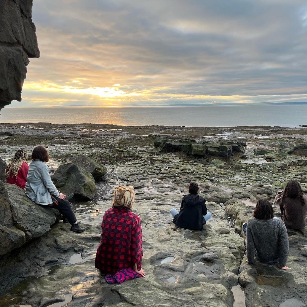 A group sits on a rocky shore watching the sunset over the ocean