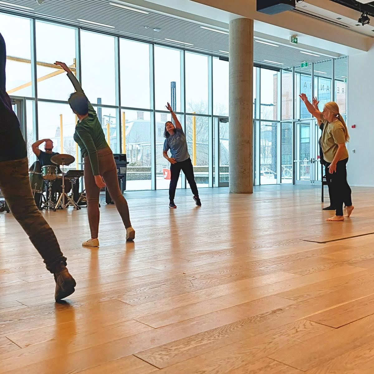 A group of people dance in a space with wood floors and a wall of windows