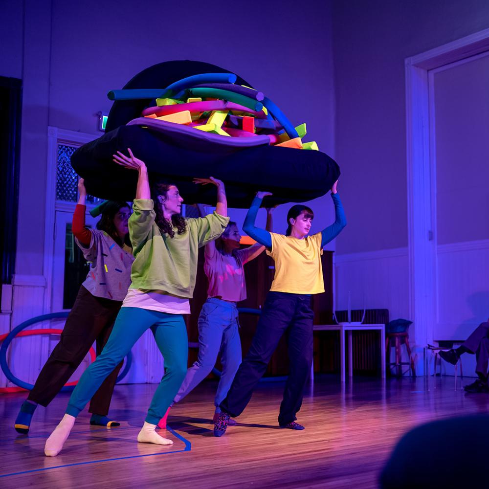 Four people hold up a raft laden with soft colourful objects.