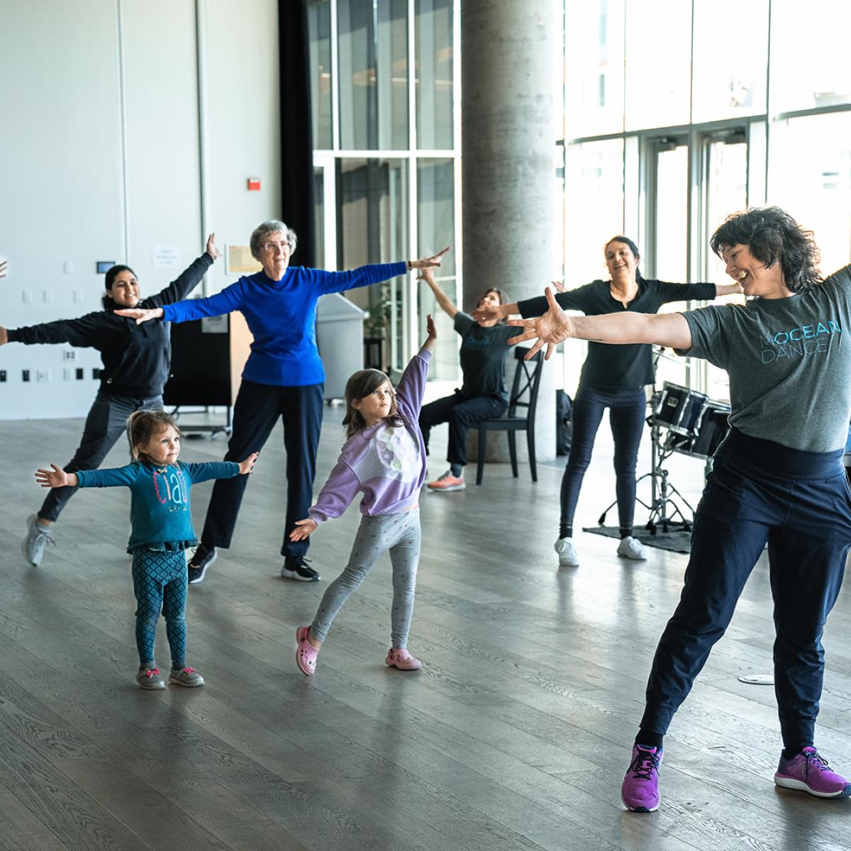 An intergenerational group stand in a well light room with arms outstretched reaching in a wide start smiling taking part in a public dance class
