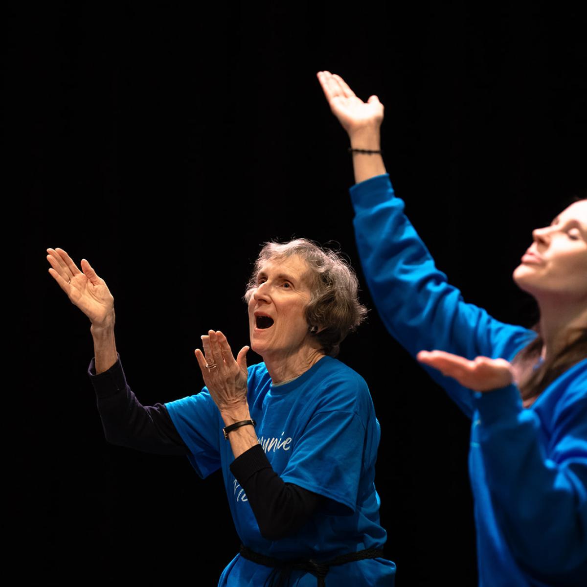 Two caucasian females stand side by side against a dark background. One female is a senior and the other late 30s, they both stand with face focused upward arms lifted in an open accepting gesture. 