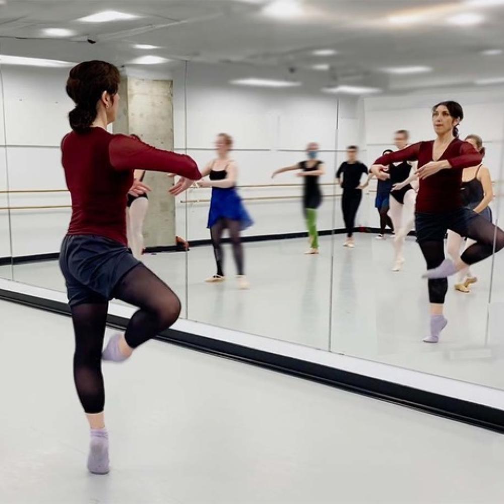 Image of a woman in a studio facing the mirror balanced on one leg in a retire position. 