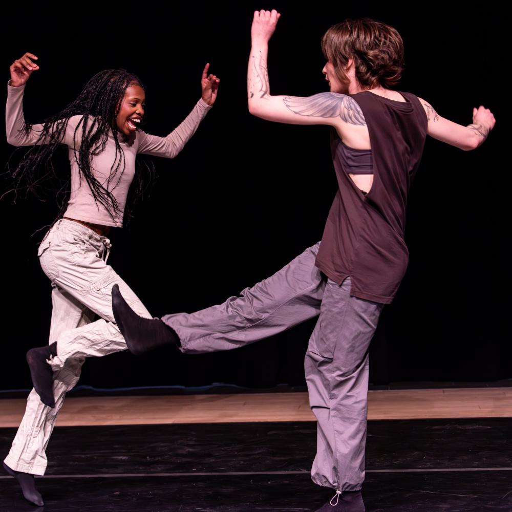 One black and one white female dancers face each other with arms in the air and hopping.