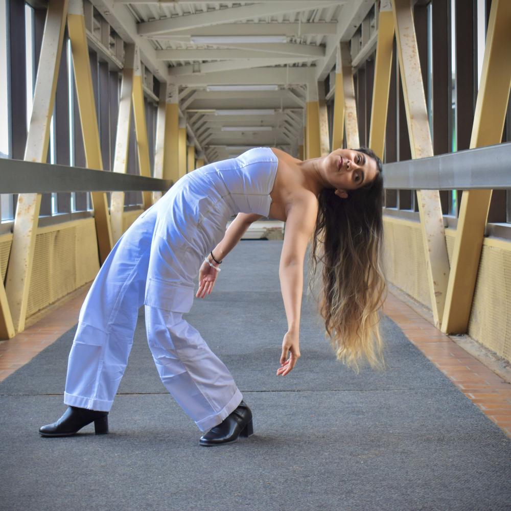 A brown female presenting dance artist with long brown hair is seen wearing a white strapless jumpsuit and black high heels. She is arched back towards the right, with her arms extended towards the ground. She is standing in a hallway with cement floors, tall windows and wooden beams