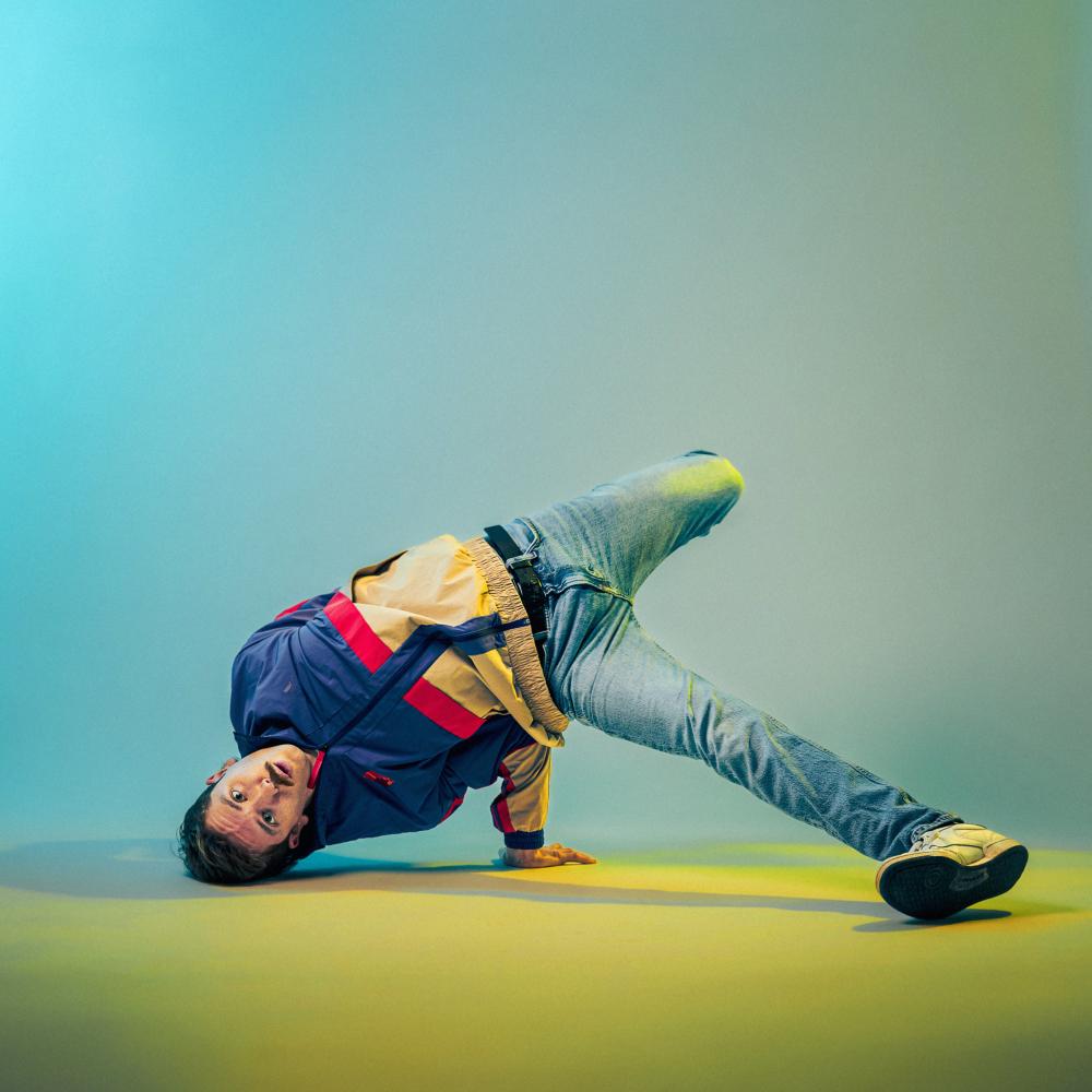A male dancer with a mustache is wearing a white, blue and red windbreaker and blue jeans. His arm, head and foot are on the ground balancing the rest of his body off of the floor. The photo is taken against a blue and yellow background.