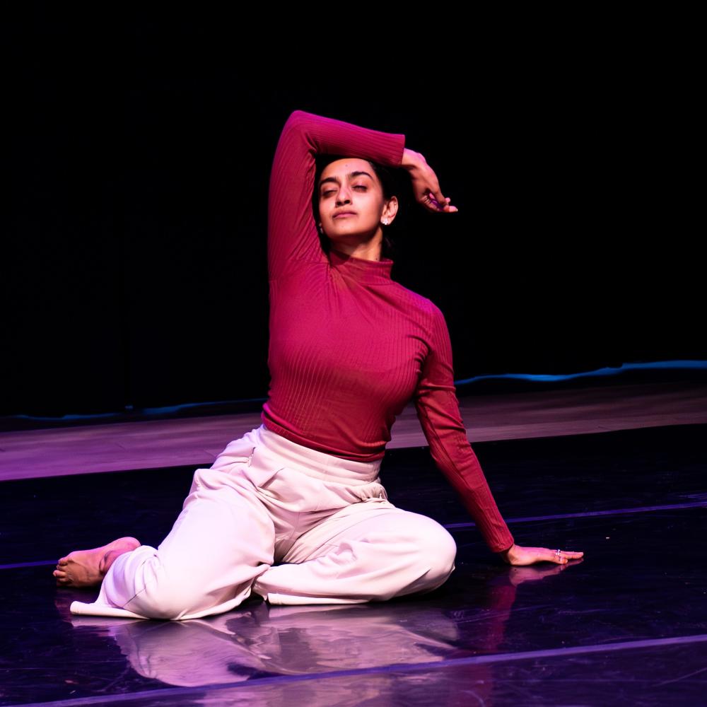 An Indian-Canadian female presenting dance artist is seen in a dark room with a black marley floor. She is wearing a red long-sleeve shirt and white pants. She is sitting on the floor, with one hand on the floor and the other arm resting on her head.