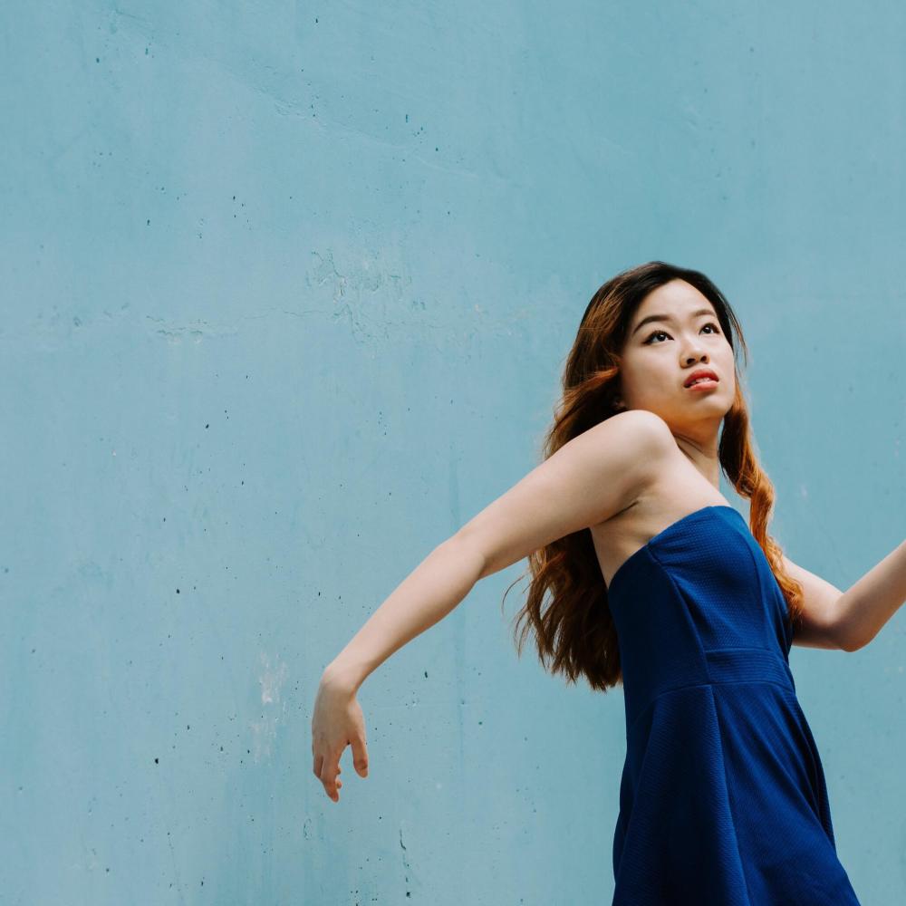 A Chinese-Canadian female presenting dance artist is seen in front of a light blue cement wall. She has long, brown, curled hair and is wearing a dark blue strapless jumpsuit. She is twisting away from the camera with curved arms