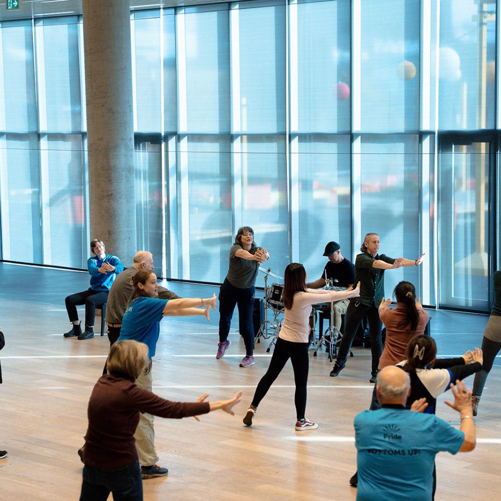 In a bright room full of large floor to ceiling windows a group of diverse people strtch to the right together in a group movement class centered around a percussionist and lead teacher wearing a shirt that says Mocean Dance. Some of the participants are seated and others standing.  