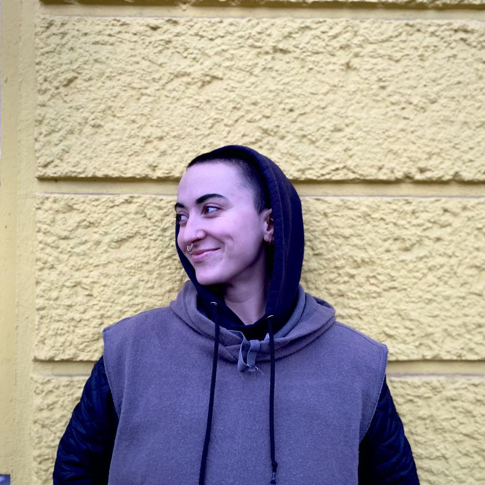 Seen against a yellow wall, a white artist with a buzz cut is standing looking to the left. They are smiling, wearing a hoodie and they have a nose ring.